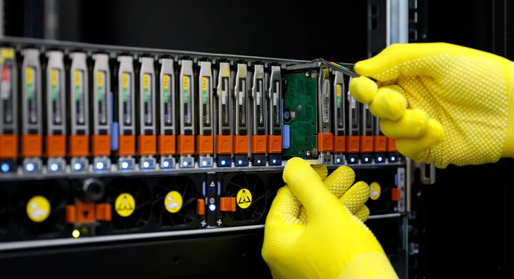 Technician wearing yellow gloves removing a hard drive from a server rack populated with multiple drives.