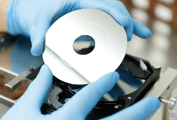 Technician wearing blue gloves carefully handling a hard disk platter during a data recovery operation in a cleanroom environment.