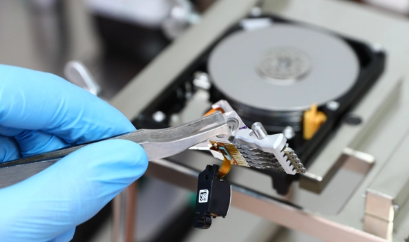 Close-up of a data recovery technician handling the read/write head assembly of a disassembled hard disk drive using precision tweezers in a cleanroom environment.