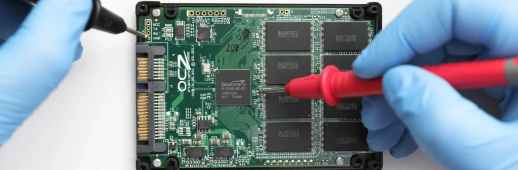 Close-up of a solid-state drive (SSD) PCB labelled "OCZ" being tested with a digital multimeter, showing hands in blue gloves probing NAND flash memory and controller circuitry for diagnostics or data recovery.